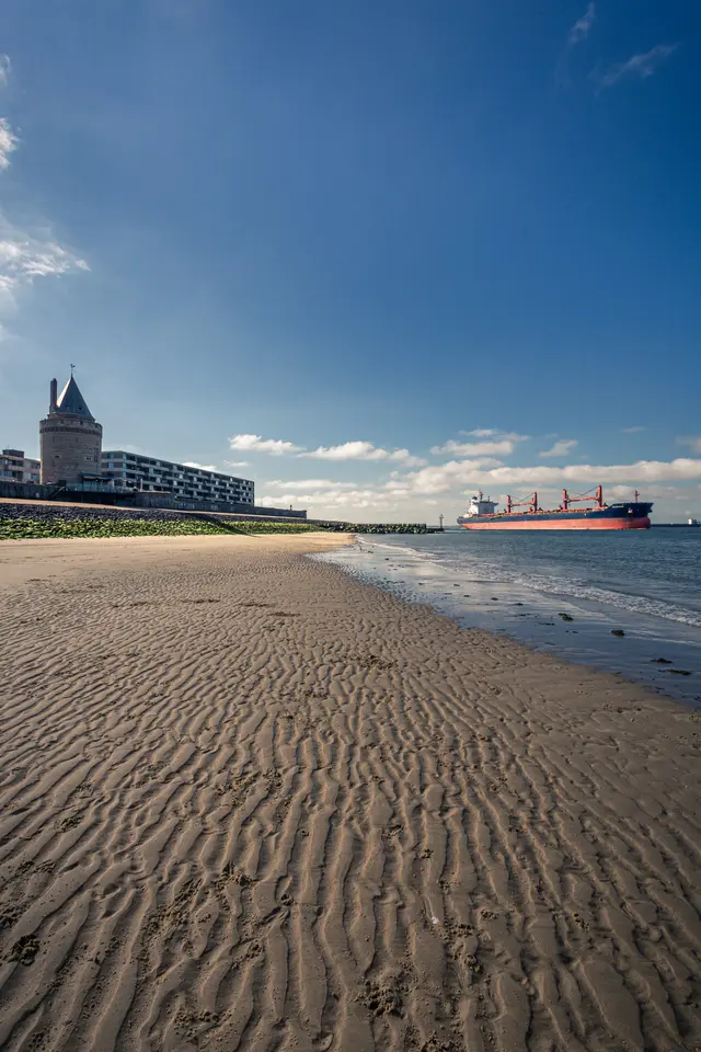 A sandy beach in Vlissingen with the Gevangentoren tower on the left and a large ship sailing in the water.