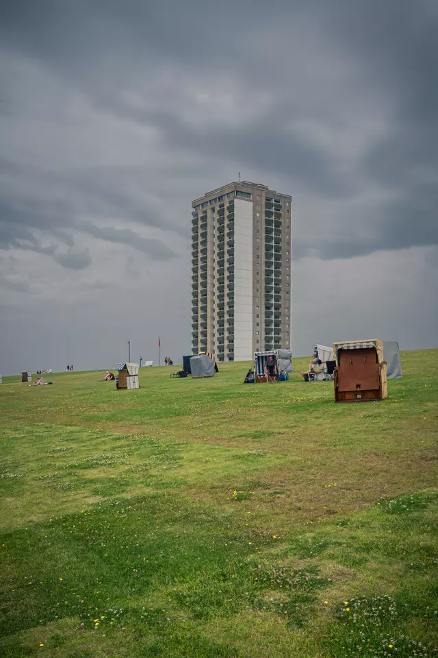 Grüne Wiese mit mehreren Strandkörben im Vordergrund und einem hohen Wohngebäude im Hintergrund unter einem bewölkten Himmel.