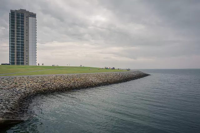 Grauer Himmel über einer Küstenlinie, links ein hoher Wohnblock und rechts ruhiges Wasser, das auf einen Kiesdamm trifft.