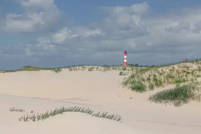 Düne mit feinem Sand, grünem Gras und einem rot-weißen Leuchtturm unter einem bewölkten Himmel.