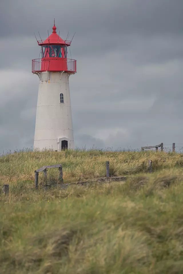 Ein Leuchtturm mit rotem Dach steht auf einer Wiese, umgeben von hohen Gräsern und einem bewölkten Himmel.