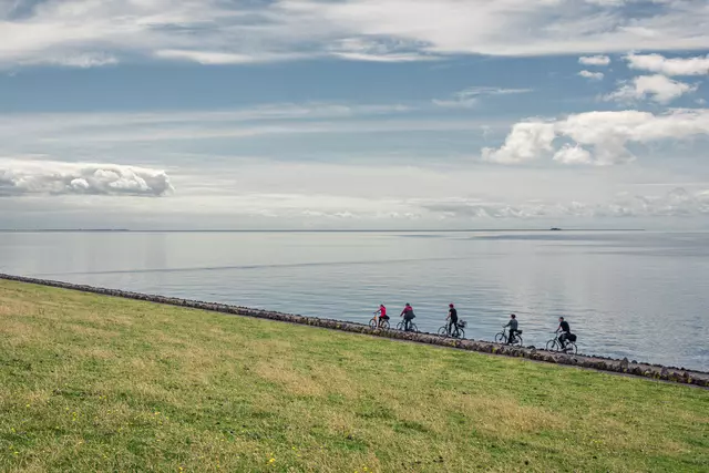 Radfahrer entlang eines Deichs mit Wasser auf der einen Seite und grüner Wiese auf der anderen. Weite Wolkenformationen am Himmel.