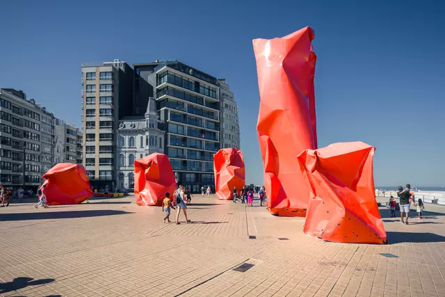 Large, red sculptures stand in a plaza surrounded by people and modern buildings under a clear sky.