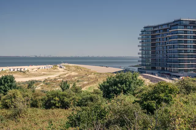 Grünflächen und ein moderner Gebäudekomplex am Wasser, mit einem Sandstrand und einer Promenade im Hintergrund.