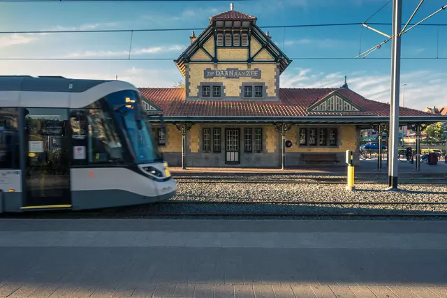 An elegant train station building with brown tones and a red roof as a modern tram passes by.