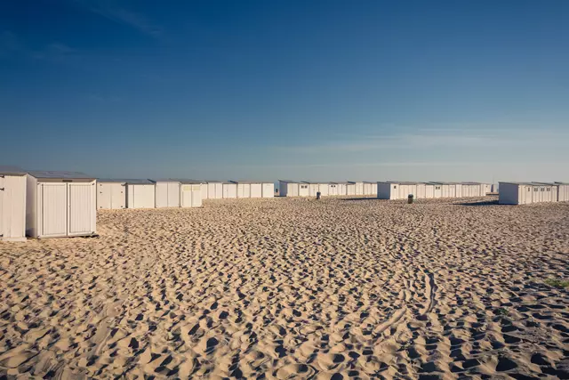 Helle Sandfläche mit mehreren weißen Strandhütten, die in einer langen Reihe aufgestellt sind, unter einem klaren blauen Himmel.