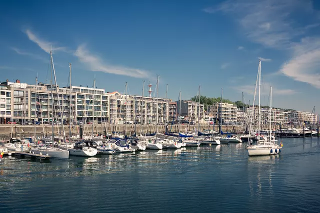 Several yachts are moored by a waterfront promenade lined with modern buildings under a clear sky.