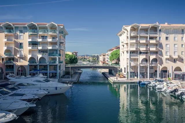 Zwei Gebäude mit Balkonen flankieren einen schmalen Kanal, in dem mehrere Boote liegen. Der Himmel ist klar und blau.