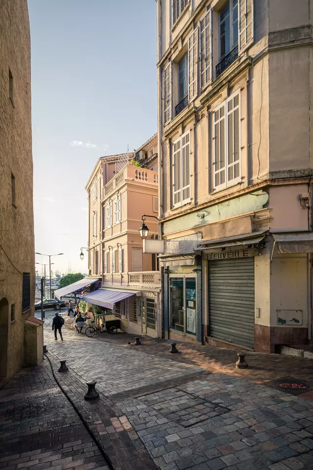 A narrow cobblestone alley surrounded by pastel buildings and a small market stall. A person walks by.